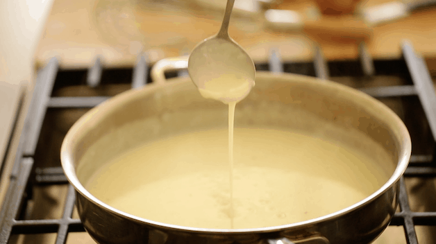 bechamel sauce in a pan on a cook top with small ladle showing the thickness of sauce