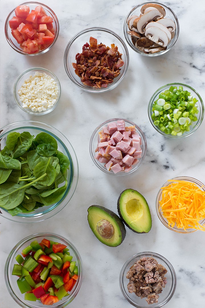 Separated ingredients that can be used in an omelet including ham, avocado, bell pepper, mushrooms, green onion, spinach, shredded cheddar cheese.