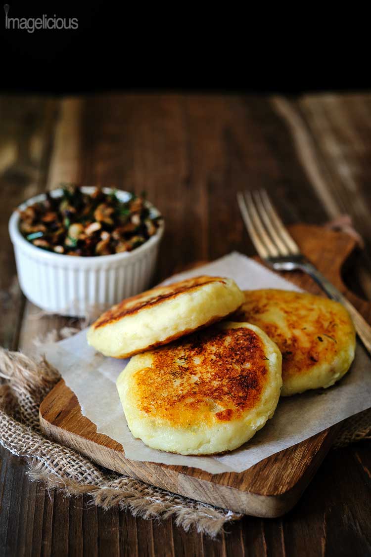 Three golden Vegan Potato Cakes stuffed with Mushrooms are on a cutting board. A bowl with mushroom stuffing is visible in the background