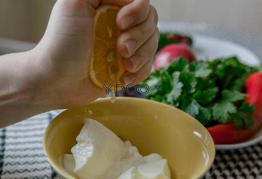 сжатая половина лимона в руке над миской с натуральным йогуртом, на фоне овощи и зелень